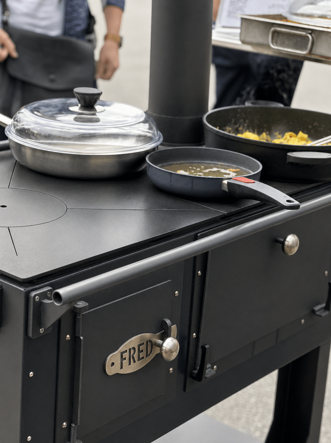 fred_schwarz2 auto_awesome Meintest du: Outdoor black grill with a frying pan cooking vegetables and a covered pot nearby; metal name plate on the side reads FRED 121 Schwarzer Außengrill mit einer Pfanne zum Braten von Gemüse und einem abgedeckten Topf daneben; ein Metallschild an der Seite trägt die Aufschrift FRED.