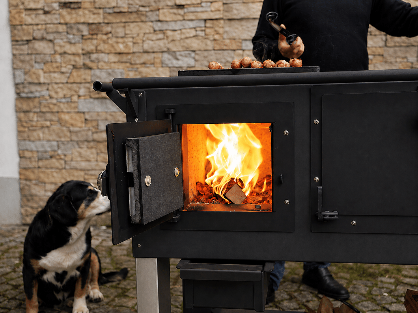 fred_schwarz3 Schwarzer Räucherofen im Freien mit offenem Feuer und Würstchen darauf; eine Person in schwarzem Hemd hält eine Grillzange, während ein Hund in der Nähe zusieht.