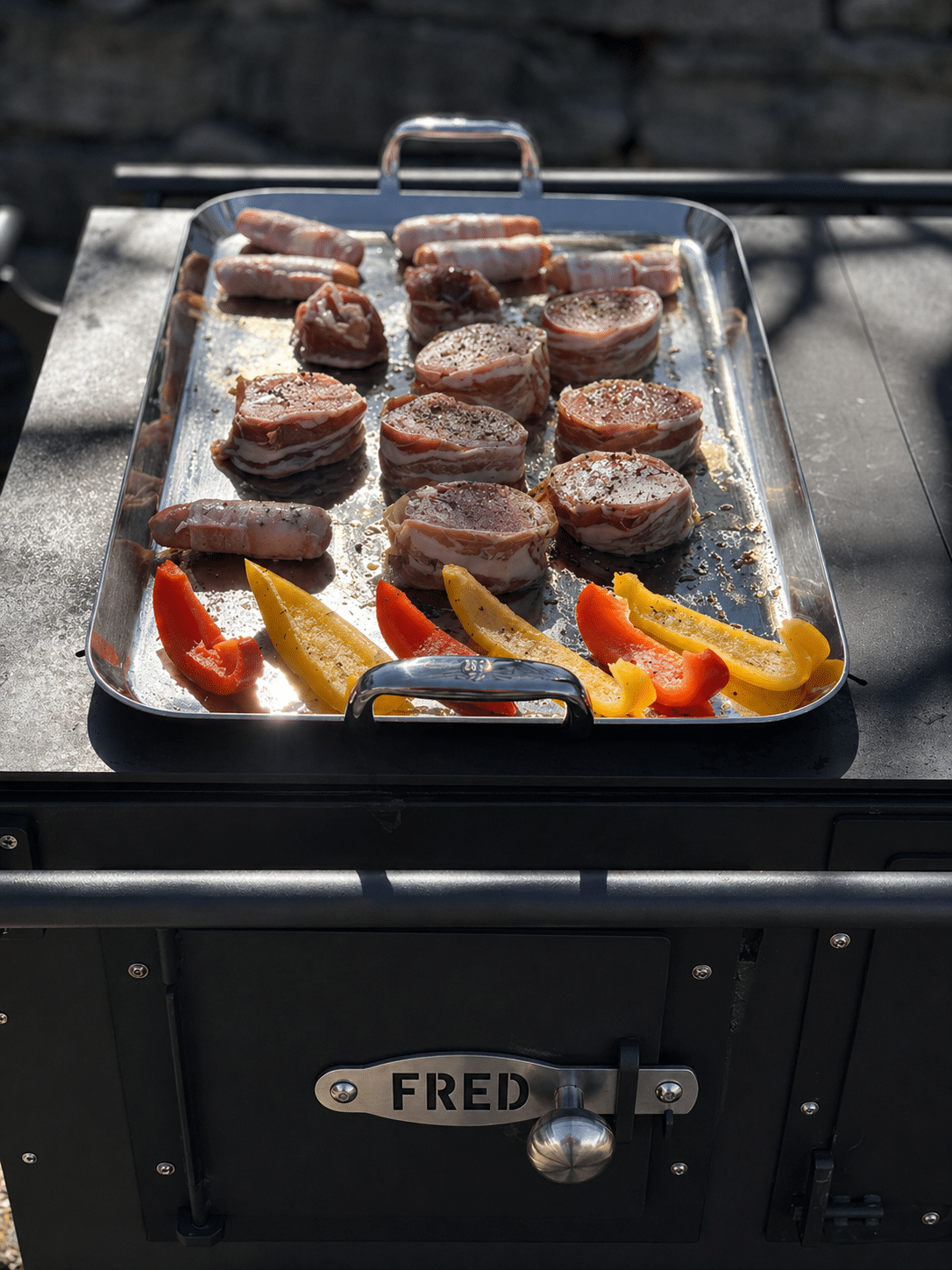 fred_schwarz4 111 Grillplatte mit in Speck gewickelten Fleischmedaillons und in Scheiben geschnittenen roten und gelben Paprikaschoten, die auf einem Außengrill garen.